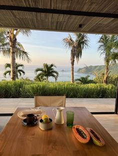 a wooden table topped with fruit next to an open patio door overlooking the ocean and palm trees