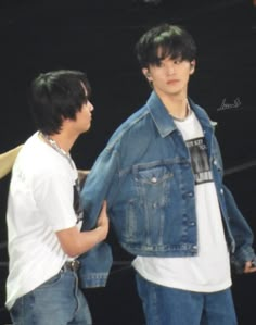 two young men standing next to each other in front of a black background, one wearing a denim jacket