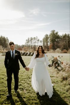 a bride and groom holding hands while walking through the grass in front of a field