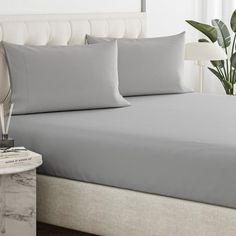 a bed with grey sheets and pillows in a bedroom next to a plant on the nightstand