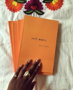 a person holding three orange books on top of a white sheet with flowers in the background