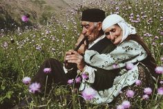 an elderly couple sitting in a field of flowers with their arms wrapped around each other