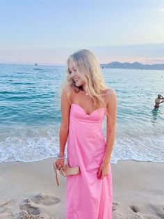 a woman in a pink dress standing on the beach with her hand in her pocket