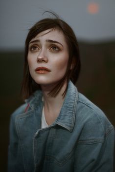 a woman with brown hair and blue jacket looking off to the side in front of a sunset