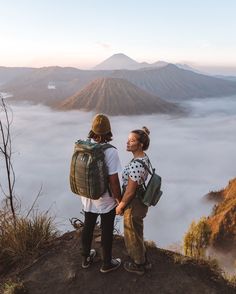 two people standing on top of a mountain looking at the clouds in the valley below