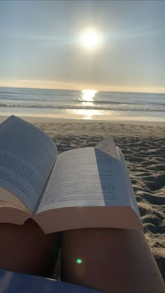 an open book sitting on top of a sandy beach next to the ocean with the sun in the background