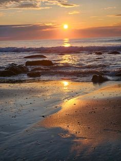 the sun is setting over the ocean with rocks in the foreground and water on the beach