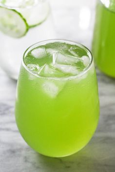 two glasses filled with limeade and ice on top of a marble table next to cucumbers