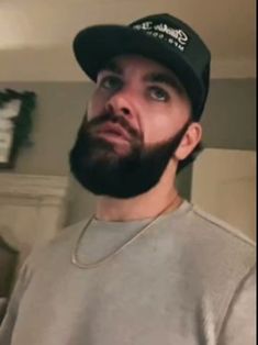 a man with a beard wearing a baseball cap and looking up at the camera while standing in a living room