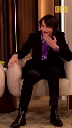 a man in a black suit and purple shirt sitting on a white chair with his hands clasped to his mouth