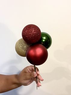 a hand is holding three christmas balls in front of a white wall with red, green and gold ornaments