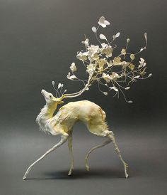 a sculpture of a deer with white flowers on it's head and legs, standing in front of a gray background