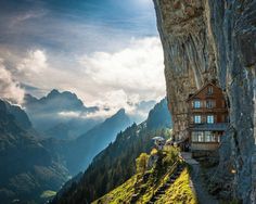 a black and white photo of a house on the side of a cliff with mountains in the background