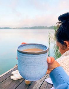 a woman holding a cup of coffee on top of a wooden deck next to the water