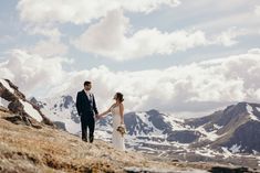 a bride and groom holding hands on top of a mountain with snow capped mountains in the background