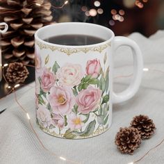 a coffee mug with pink roses on it next to pine cones and christmas lights in the background