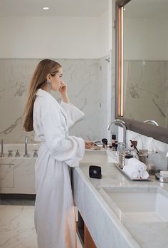 a woman in a bathrobe is standing by the bathroom sink talking on her cell phone