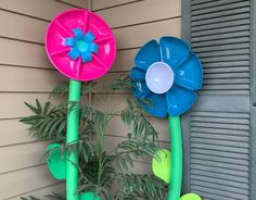 two plastic flowers are next to each other on the side of a house with green plants