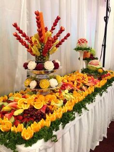 an arrangement of fruits and flowers on a table with white drapes in the background