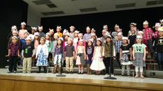 a group of children standing on top of a stage wearing paper hats and costumes with words written in front of them