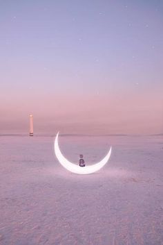 a person sitting on top of a crescent moon in the middle of a field with a lighthouse in the background