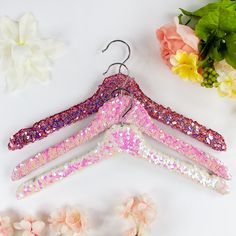 three pink and purple glittered clothes hangers next to flowers on a white surface