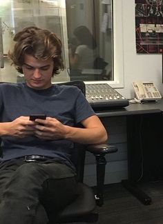 a young man sitting in a chair looking at his cell phone while he works on the computer