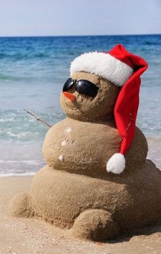 a sand snowman wearing sunglasses and a santa hat on the beach with blue water in the background