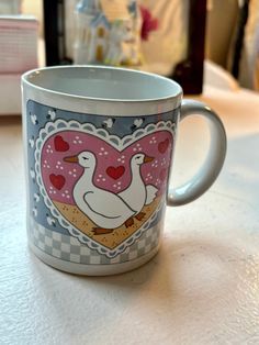 a coffee cup with two geese on it sitting on a table next to a window