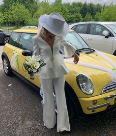 a woman standing next to a yellow car wearing a white suit and hat with flowers in her hand