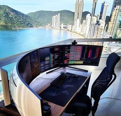 a computer desk with multiple monitors on it in front of a large window overlooking the ocean