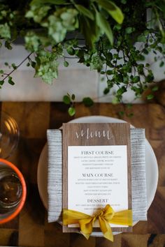 a place setting with a menu and yellow ribbon on the napkin, next to a glass vase filled with greenery