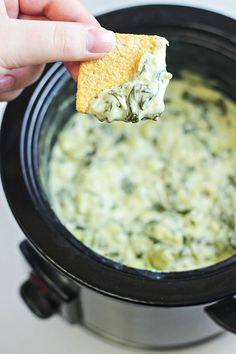 a hand holding a piece of bread over a bowl of spinach and artichoke dip