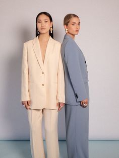 two women standing next to each other in front of a white wall wearing suits and pants