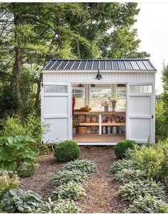 a small white shed in the middle of some trees and bushes with potted plants