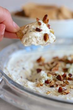 a hand holding a cracker over a bowl of cream cheese with nuts on top