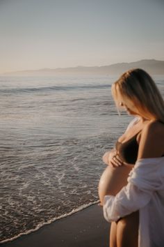 a pregnant woman sitting on the beach next to the ocean with her stomach exposed and looking at the water