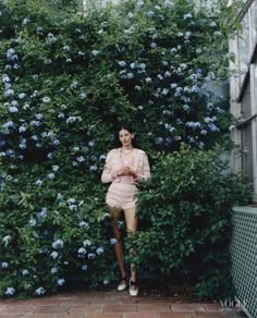 a woman standing in front of a bush with blue flowers on the bushes behind her