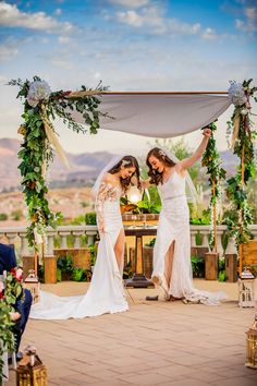 two women in wedding dresses standing next to each other under an arch covered with greenery