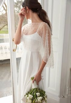 a woman in a white dress is holding a flower bouquet and looking out the window