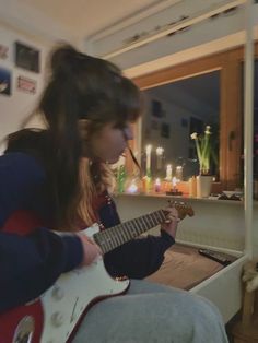 a young woman playing an electric guitar in her living room with candles on the window sill