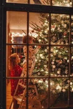 two children are decorating a christmas tree through a window with lights in the background