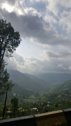 a bench sitting on top of a lush green hillside under a cloudy sky with mountains in the distance