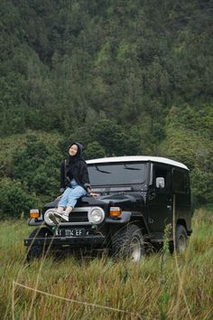 a man sitting on the hood of a black jeep in tall grass with trees in the background