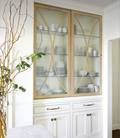 a white china cabinet with glass doors and plates on it's sides, next to a potted plant