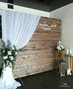 a wooden wall with white drapes and flowers on the floor next to a table