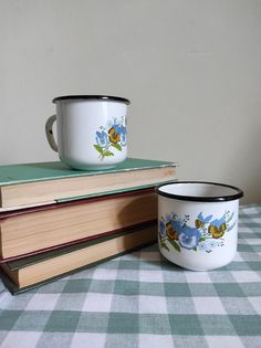 two coffee mugs are sitting on top of some books and one has a flower painted on it