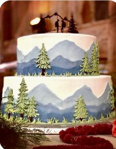 a three tiered cake with trees and mountains painted on the side, surrounded by greenery