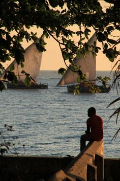 a man sitting on a wall looking out at the ocean with sailboats in the distance