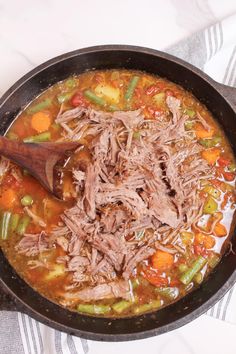 a skillet filled with meat and vegetables on top of a white table cloth next to a wooden spoon